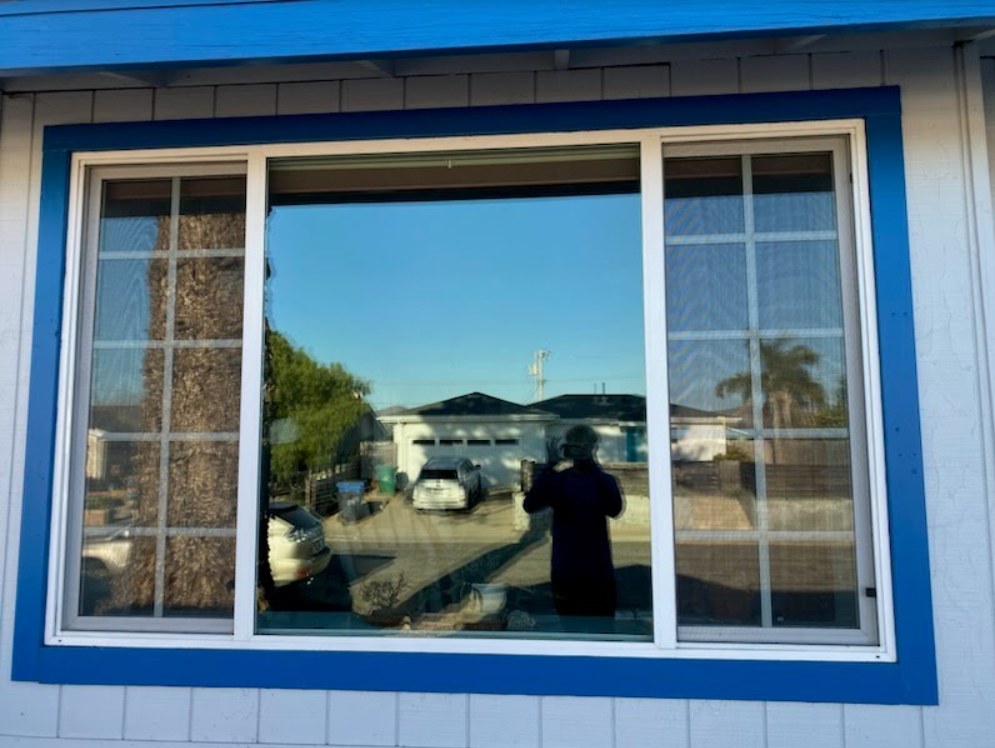 Large picture window with blue trim flanked by two screened panes, the glass clear enough to reflect a driveway and blue sky